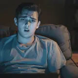 Young man on a couch in a dim living room illuminated by blue device glow, looking confused and offended