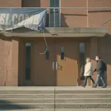 Torn church welcome banner above entrance doors with frayed edges and two visitors approaching