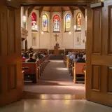 View through open church doors toward a distant pastor at the pulpit, warm stained glass light filling the sanctuary