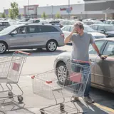 Frustrated man pinching the bridge of his nose beside his car in a crowded retail parking lot at midday