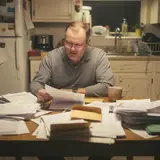 Middle-aged man at kitchen table surrounded by stacks of bank statements with a bewildered expression